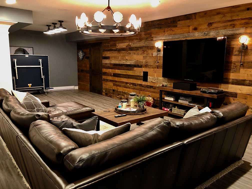 High end luxurious basement remodel with a wood accent wall, a wall mounted TV, a leather sectional couch, and a decorative chandelier