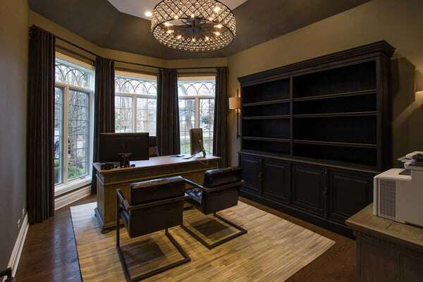 Home office with a decorative chandelier above two meeting chairs and a desk with 3 large floor to ceiling windows behind