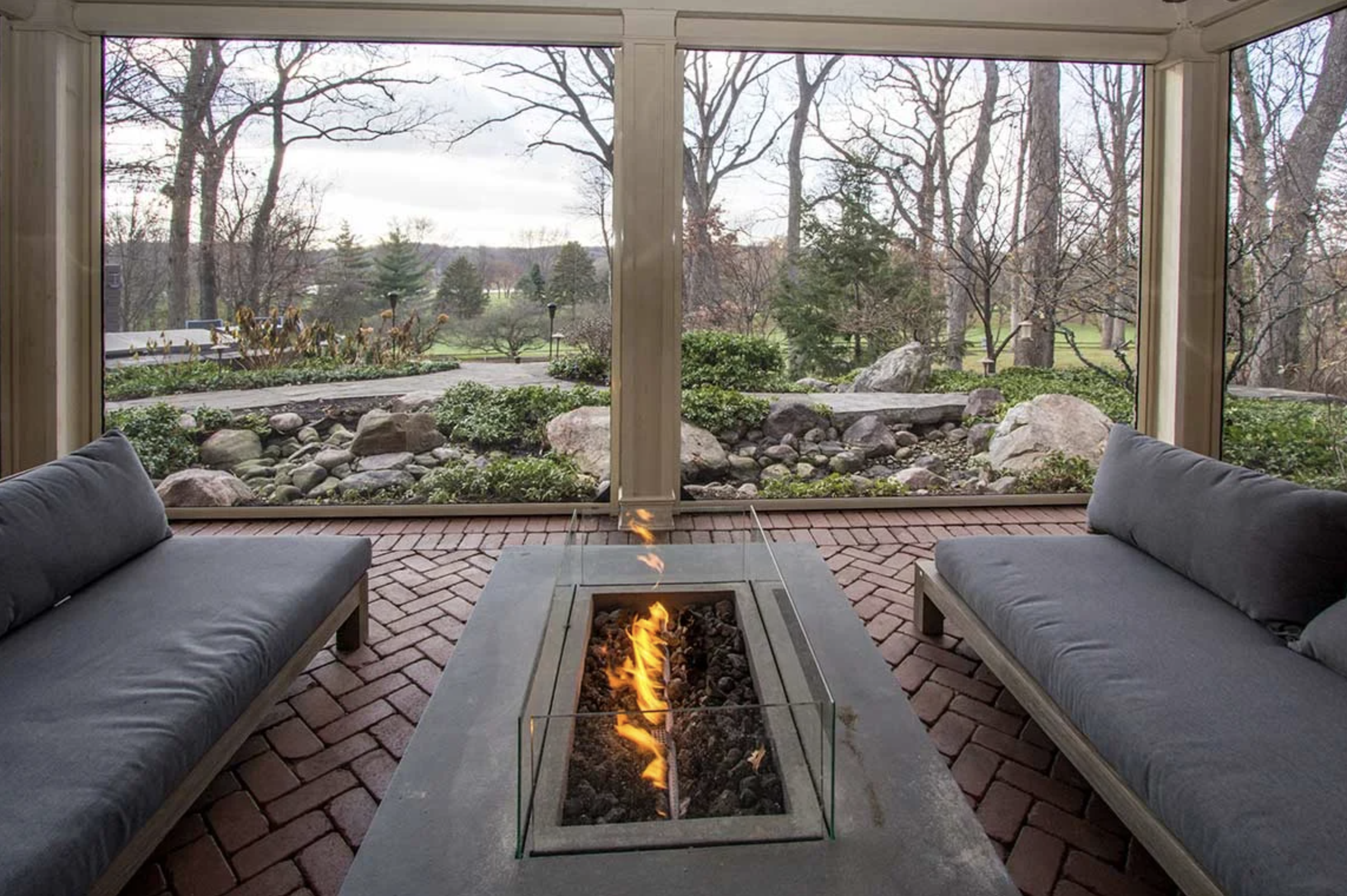 Outdoor screened in porch with a parralleled seating area and a firepit built into the table top placed between the armless loveseats