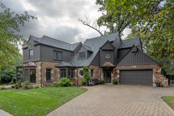 Street view photo of a vintage home with brick walls and black exterior panelling. There is a brick driveway that leads up to a two car garage and a two story home